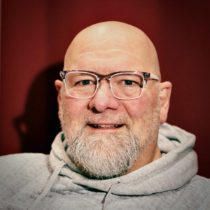 A bald, older White man with a grey beard, smiling. Wearing glasses and a grey hoodie.